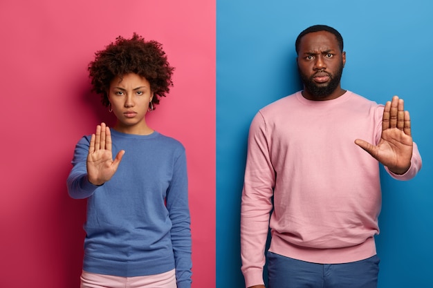 Символ запрета. Серьезные недовольные темнокожие мужчина и женщина делают стоп-жест ладонями, недовольно смотрят, носят повседневную одежду.