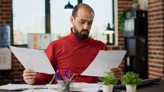 Project manager analyzing e commerce information on files, looking at documents and computer screen. Male employee working on financial analysis with statistics, charts and graphs.