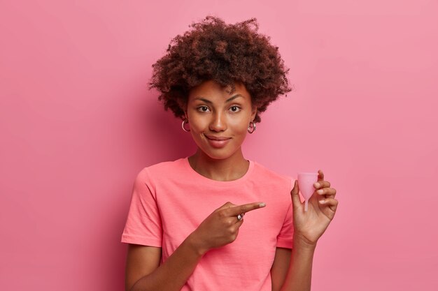 Protection for female hygiene. Afro American woman points at reusable menstrual cup, demonstrates product for menstruation, best way of protection, good alternative, isolated on pink wall