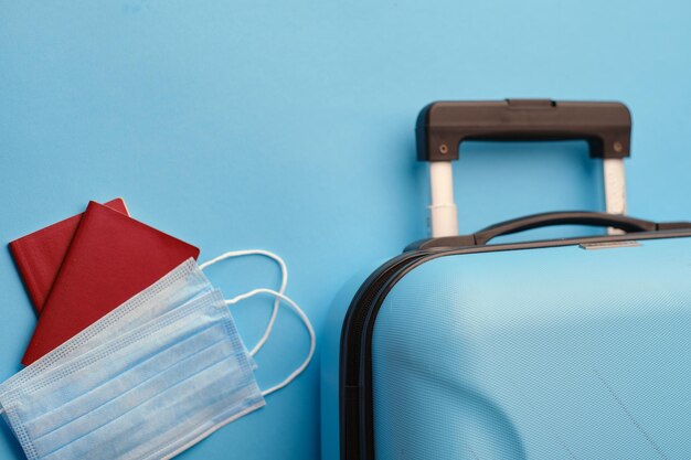 Protective Medical face mask and passport with suitcase on blue background top view