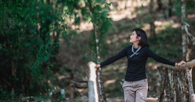 Выдающаяся молодая женщина, стоящая на бамбуковом мосту, улыбается и смотрит на красивую природу во время кемпинга в лесу со счастливым местом для копирования