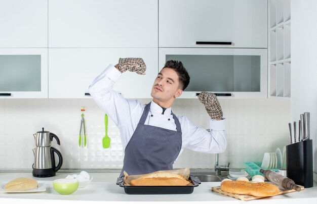 Proud and ambitious male commis chef in uniform wearing holder and freshly-baked bread in the white kitchen