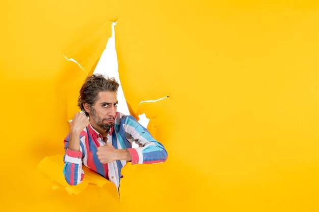 Free photo proud and ambitious young guy posing for camera through a torn hole in yellow paper