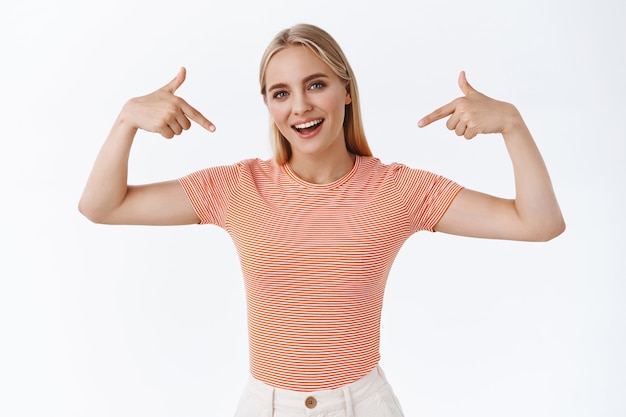 Proud, boastful good-looking stylish young woman with blond hair, tattoos, standing striped t-shirt, pointing herself as bragging, smiling sassy, show-off as gain promotion, white background