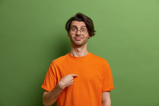 Proud cheerful man points at himself and asks who me, has pleased expression, dressed in bright orange t shirt, round transparent glasses, isolated on green wall, boasts about his achievements