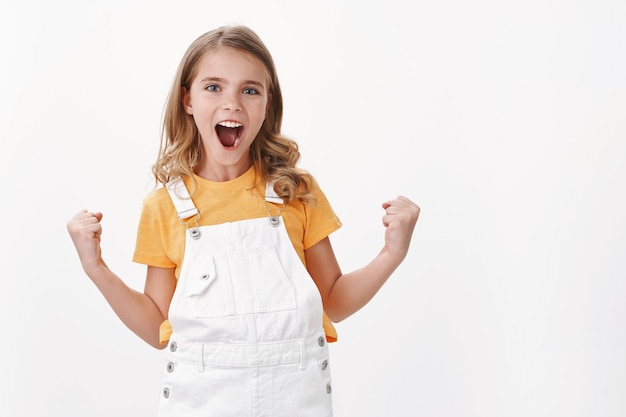 Proud good-looking young satisfied little girl with fair hair and blue eyes, fist pump celebration gesture, smiling joyfully, achieve success, triumphing from good excellent news, white wall