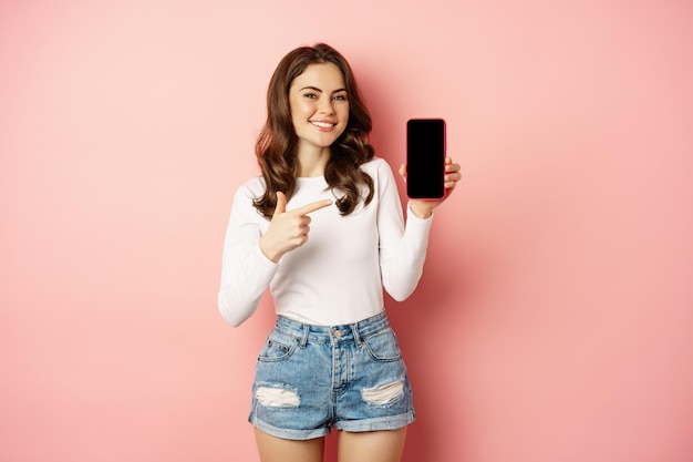 ピンクの背景の上に立って、スマートフォンで携帯電話の画面、携帯電話のアプリまたはプロモーションテキストを表示、人差し指の誇りと幸せな笑顔の女の子