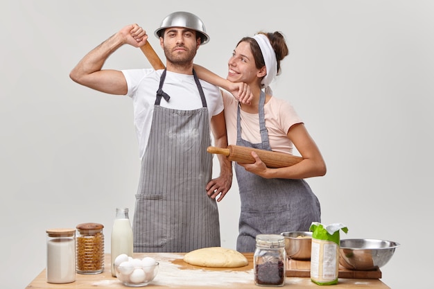 Proud man and his cheerful wife busy at kitchen, dressed in aprons, finish making dough, bake bread together, use secret ingredient, stand against white wall near table with fresh products
