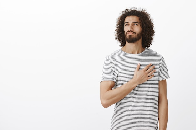 Orgoglioso del mio paese. ritratto di uomo orientale thoutful bello con barba e capelli ricci, tenendo il palmo sul cuore e distogliere lo sguardo con appassionata espressione premurosa