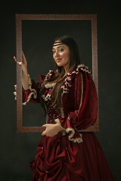 誇りに思う。暗い背景の上に立っている赤い古着の中世の若い女性の肖像画。公爵夫人、王室の人としての女性モデル。時代、現代、ファッション、美しさの比較の概念。