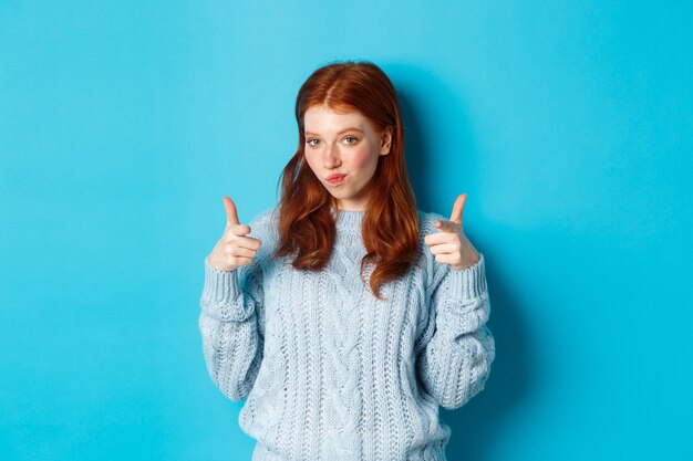 Proud redhead girl pointing fingers at camera, praising and saying congrats, standing over blue background. Copy space