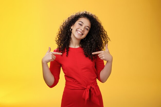 Proud and satisfied ambitious successful female student in red dress standing pleased smiling and pointing at herself as if bragging about own achievements happily and glad over yellow wall.