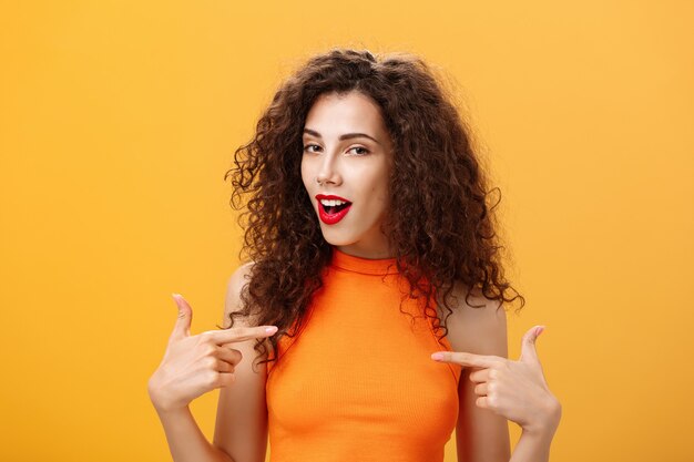 Proud and satisfied cool urban female with red lipstick and curly hairstyle pointing at herself with self-assured expression winking bragging about skills and achievements over orange background.