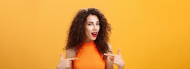 Proud and satisfied cool urban female with red lipstick and curly hairstyle pointing at herself with