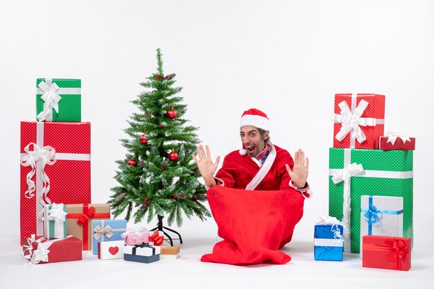 贈り物と白い背景の上の地面に座って飾られたクリスマスツリーでサンタクロースに扮した誇り高き若い大人