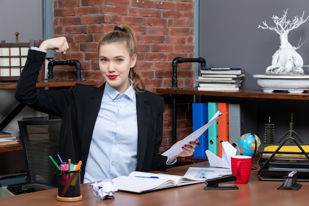 Гордая девушка сидит за столом и держит в руках документ, показывающий ее силу в офисе