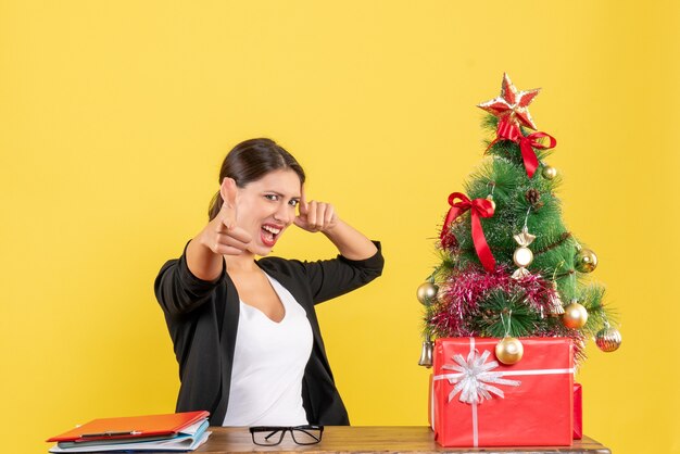 Proud young woman in suit with decorated Christmas tree at office on yellow 