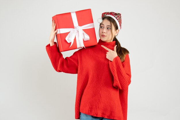 prying girl with santa hat holding present finger pointing her present standing on white