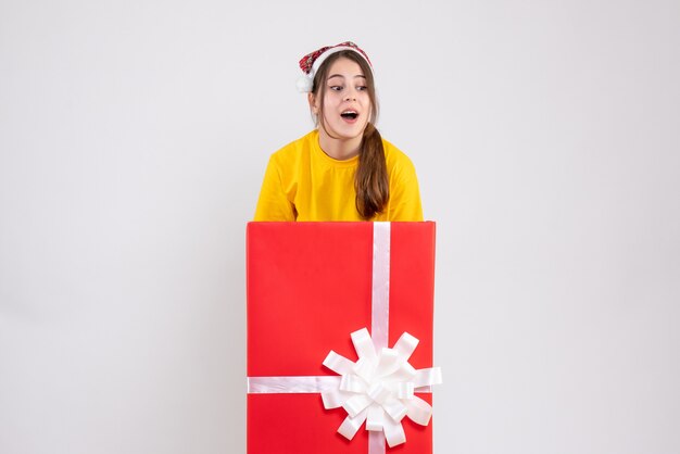 白の大きなクリスマスプレゼントの後ろに立っているサンタ帽子を持つ詮索好きな女の子