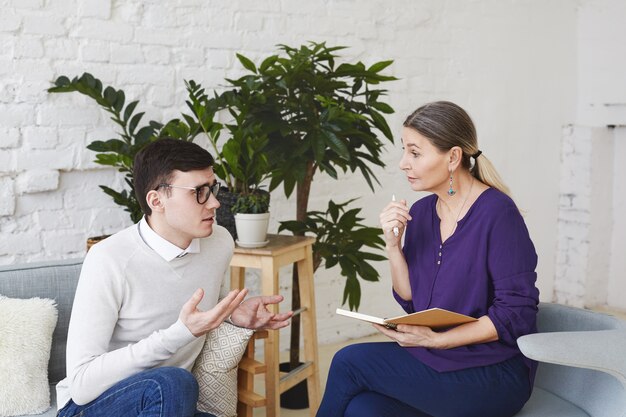 Psychology, therapy, psychiatry, mental health and counseling concept. Candid shot of nervous self conscious young male in glasses telling middle aged female counselor about his problems at work