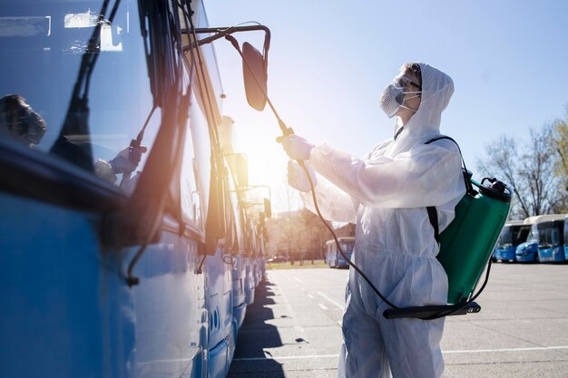 Public transport disinfection Man in white protective suit with reservoir spraying disinfectant on parked buses. Stop coronavirus or COVID-19.