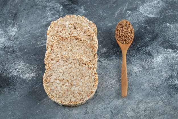 Puffed crispbread and wooden spoon of raw buckwheat on marble surface