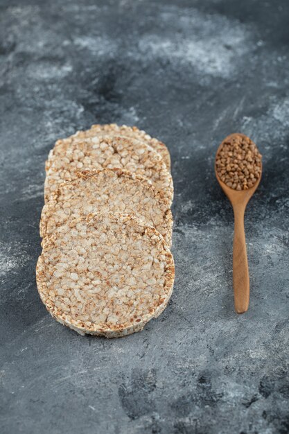 Puffed crispbread and wooden spoon of raw buckwheat on marble surface