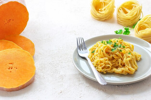 Тыквенные макароны альфредо феттуцин в керамической тарелке со свежими сырыми кусочками тыквы. Осенняя еда на обед. Рецепт тыквенного ореха.