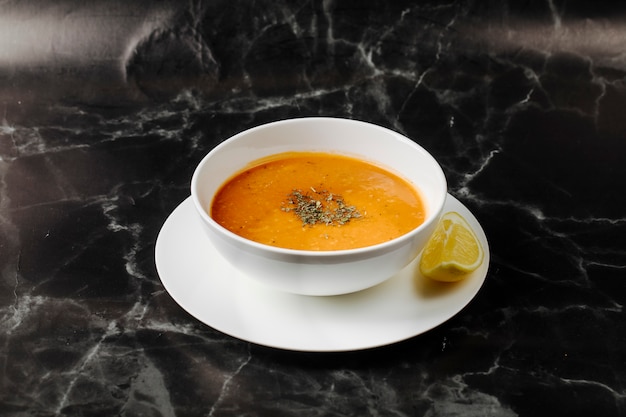 Pumpkin soup inside white bowl with herbs and spices on it with a slice of lemon around.