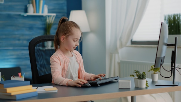 책상에서 온라인 수업 수업을 위해 키보드와 컴퓨터를 사용하는 학생. 학교 과제와 숙제를 위해 모니터를 보고 있는 어린 소녀. 원격 교육 및 지식을 위해 PC에서 작업하는 어린 아이