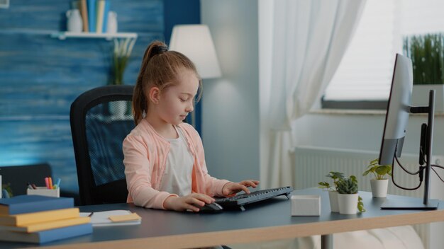 Pupil using keyboard and computer for online class lessons at desk. Young girl looking at monitor for school task and homework. Little child working on pc for remote education and knowledge