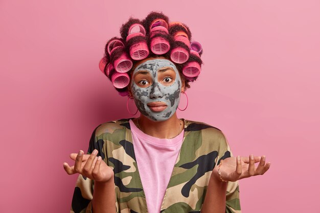 Pure beauty and hairstyling concept. Doubtful woman with hair curlers and facial mask, spreads palms sideways, doesnt know answer on difficult question, dressed in domestic clothes, isolated on pink