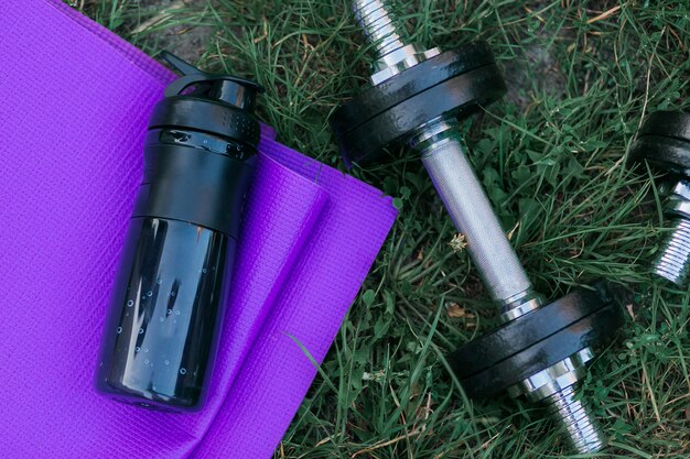 purple yoga mat, water bottle and black dumbbell on green grass. 