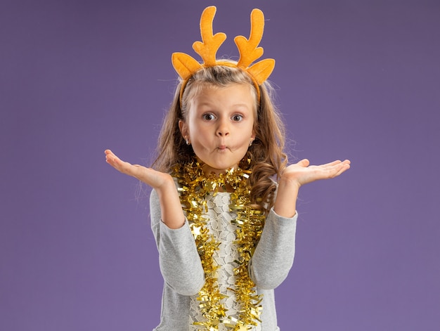 Pursing lips little girl wearing christmas hair hoop with garland on neck spreading hands isolated on blue background