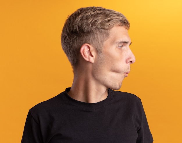 Pursing lips looking at side young handsome guy wearing black shirt isolated on yellow wall