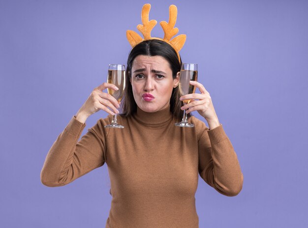 Pursing lips young beautiful girl wearing brown sweater with christmas hair hoop holding two glass of champagne around ears isolated on blue background
