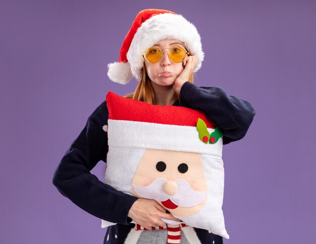 pursing lips young beautiful girl wearing christmas sweater and hat with glasses holding christmas pillow putting hand on cheek isolated on purple wall