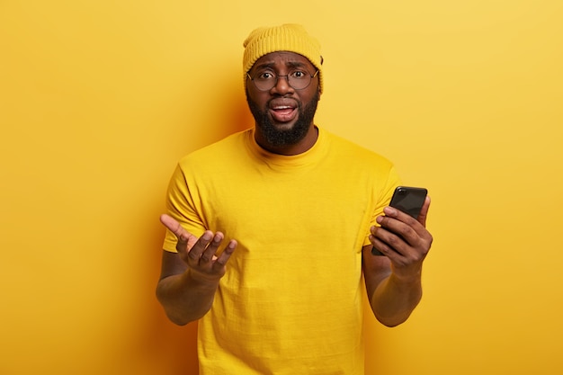 Puzzled Afro American man looks with frustrated expression, holds modern cellular