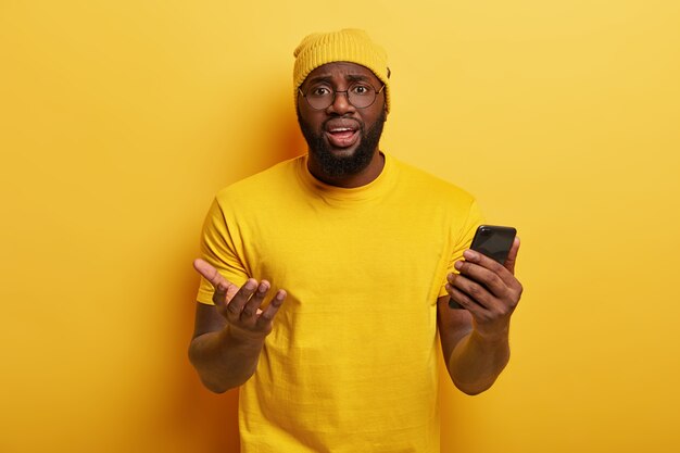 Puzzled Afro American man looks with frustrated expression, holds modern cellular