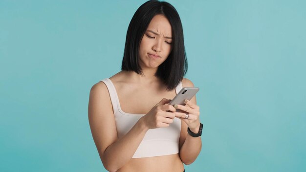 困惑したアジアの女性がスマートフォンを使用してオンラインで商品を選択し、青い背景で隔離現代の技術