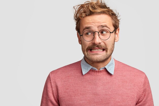 Puzzled attractive bearded emotive man wonk in spectacles, looks with worried embarrassed expression aside