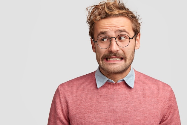 Puzzled attractive bearded emotive man wonk in spectacles, looks with worried embarrassed expression aside