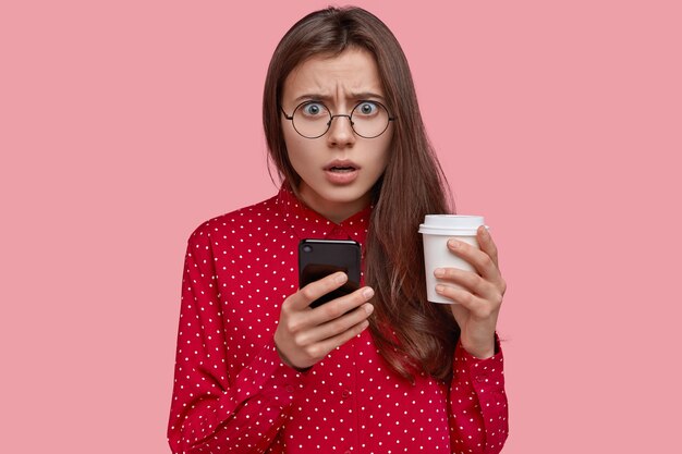 Puzzled attractive young lady with long straight hair, holds modern mobile phone, drinks takeaway coffee, disappointed with weird message, wears round glasses