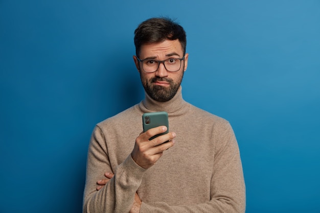 Foto gratuita ragazzo barbuto perplesso con gli occhiali utilizza un moderno telefono cellulare