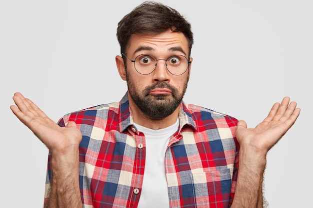 Puzzled bearded young man looks with surprised and hesitant expression, shrugs shoulders in bewildermentindoor