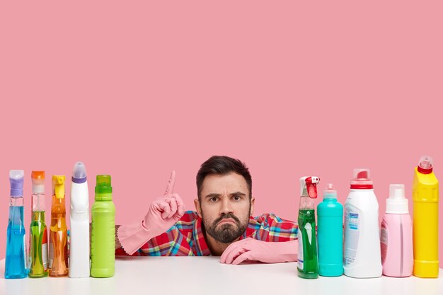 Puzzled bearded young man points upwards, has displeased expression, shows place to clean, uses high quality detergents