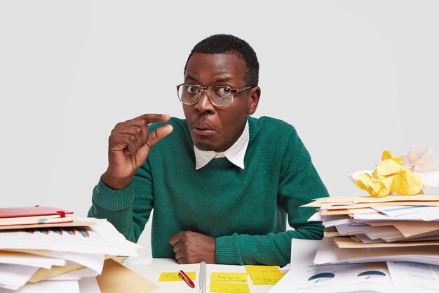 Puzzled black African American guy with short hair, makes little gesture, has confused facial expression, demonstrates how much time has for finishing work