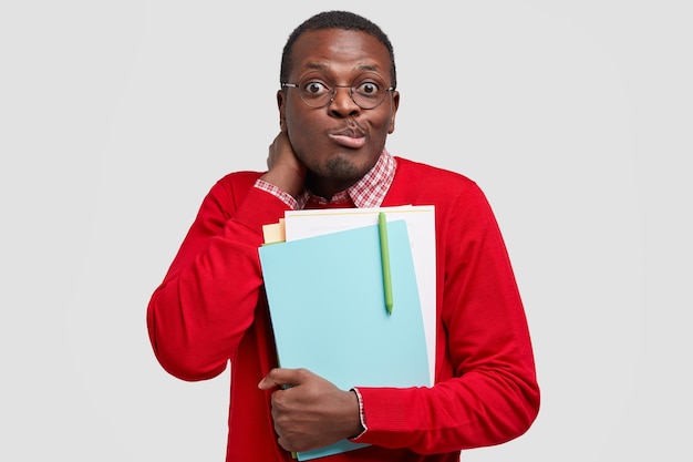 Puzzled black ethnic schoolboy purses lips with bewilderment, carries papers and textbooks closely, wears red clothes