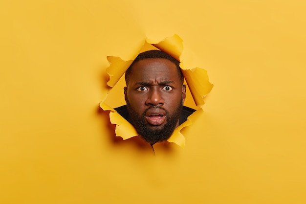 Free photo puzzled black man with thiсk bristle, looks with angry surprised face expression, keeps head in torn paper hole, stands bothered and disappointed. yellow background