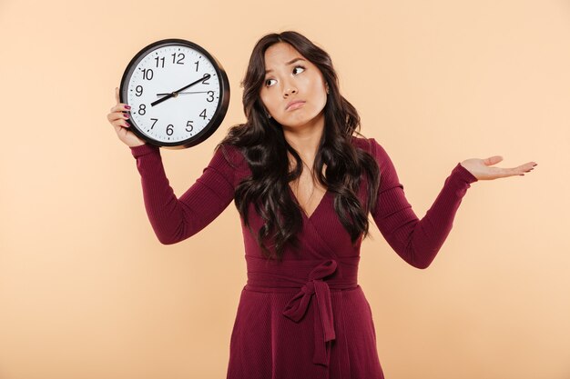 Puzzled brunette woman with curly long hair holding clock showing time after 8 gesturing like she is late or do not care over peach background